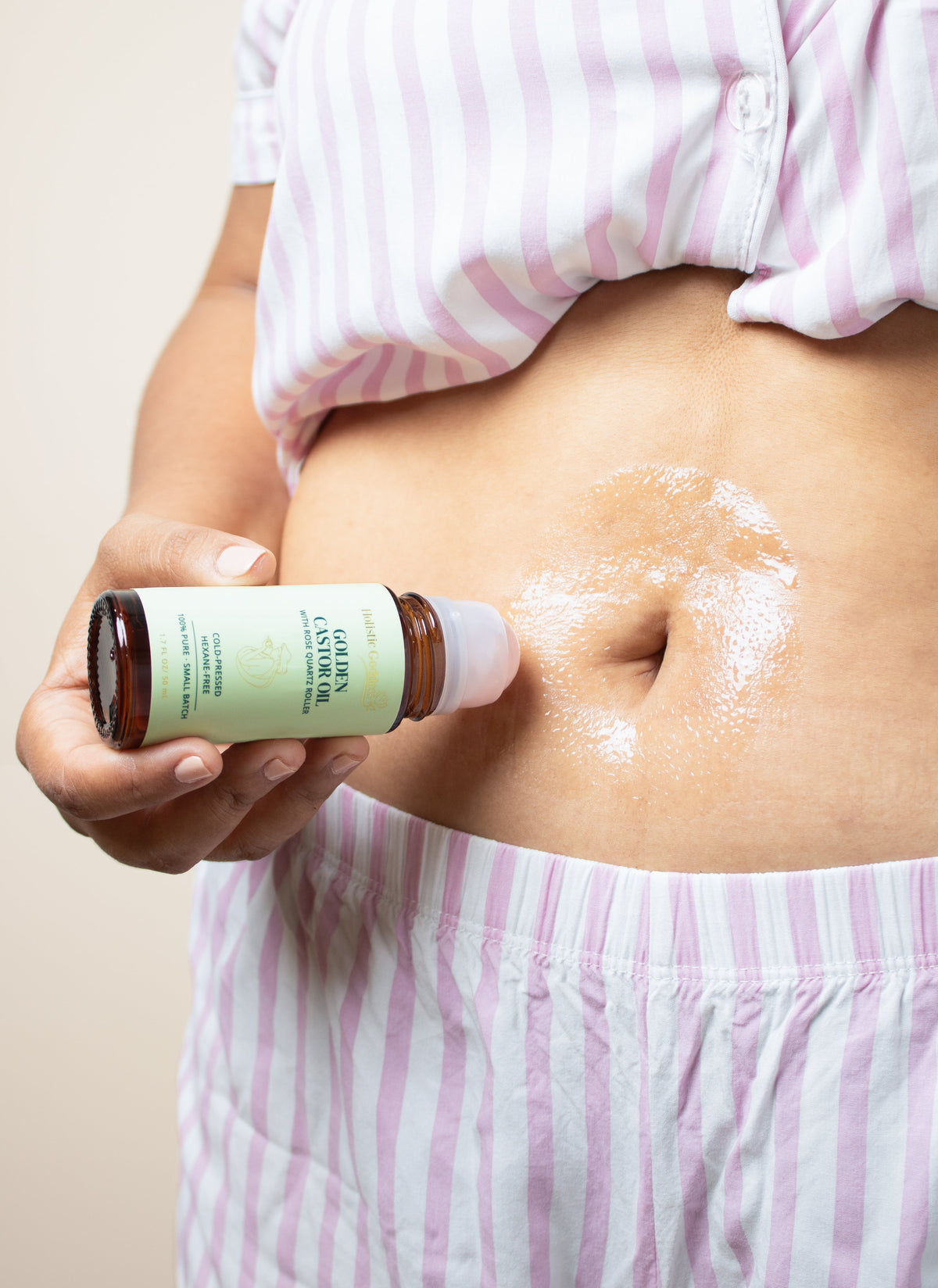 Woman applying Golden Castor Oil roller to belly, close-up lifestyle photo in pink striped pajamas