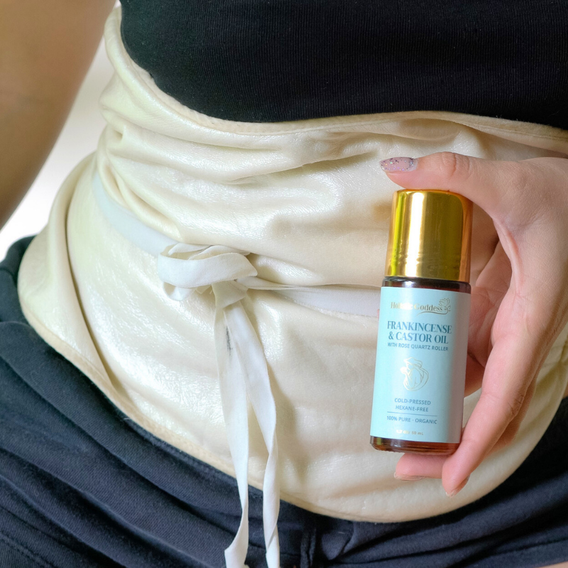 Hand holding the Frankincense & Castor Oil roll-on in front of a castor oil pack tied at the waist
