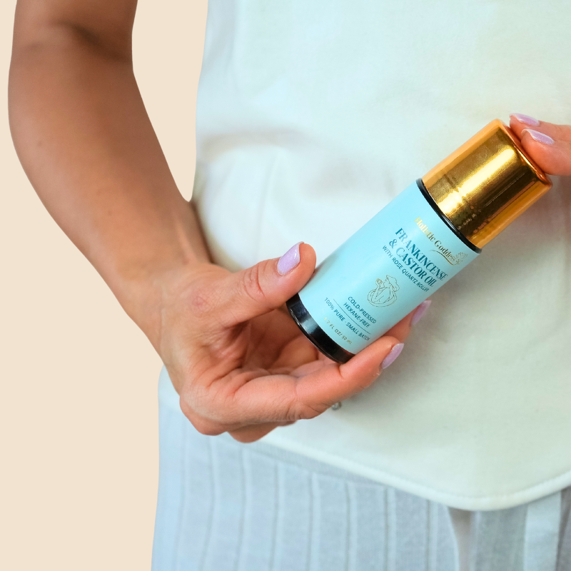 Hand holding the Frankincense & Castor Oil roll-on in front of a woman wearing a castor oil pack