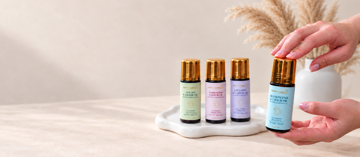Four castor oil bottles with gold caps displayed on a white tray, with a person's hands holding one the Frankincense Castor Oil next to them on a beige tabletop.