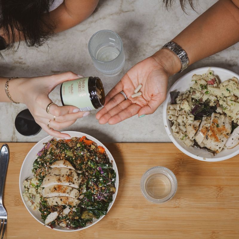 WellBelly – overhead shot of two people pouring capsules into hand at table set with salad bowls, lifestyle photo