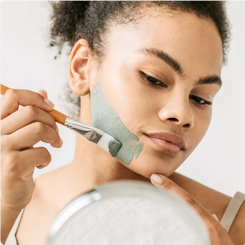 Bentonite Volcanic Clay by Yareli Wellness – woman applying clay mask with brush, looking in mirror